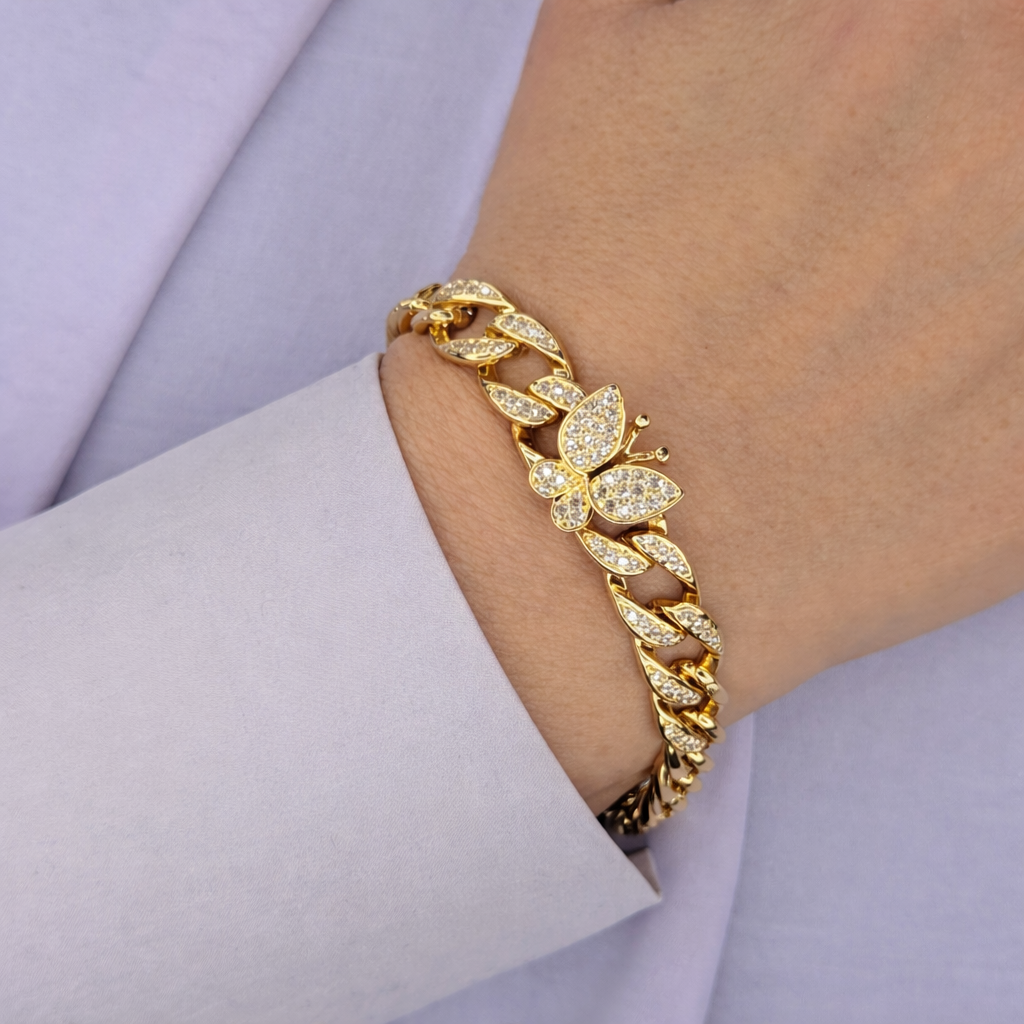 Gouden armband met schakelontwerp en vlinderdetail bezet met fonkelende stenen, gedragen om de pols tegen een lichte achtergrond.