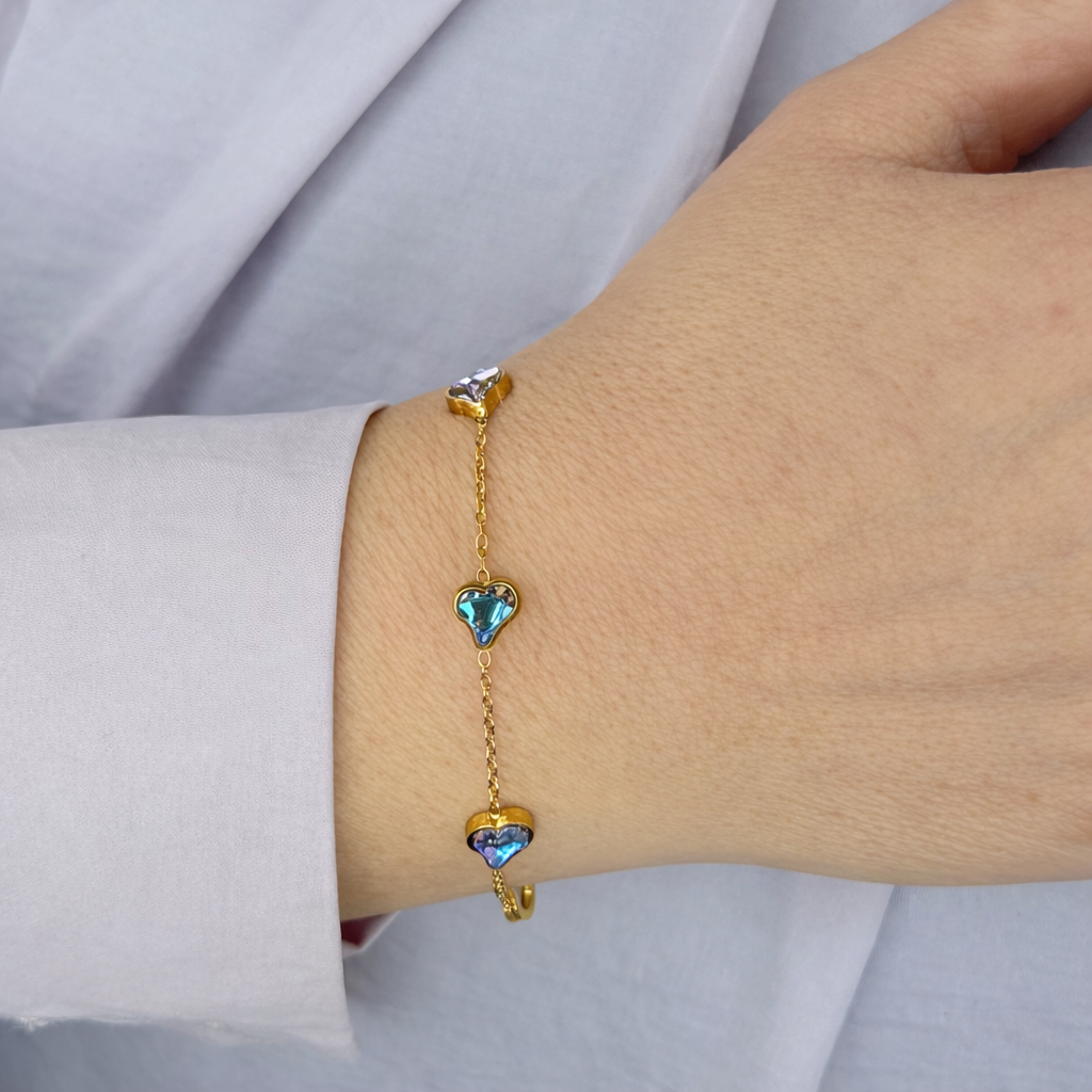 Gouden armband met fijne schakel en drie hartvormige stenen in blauwgroene tinten, gedragen om de pols tegen een lichte achtergrond.