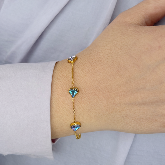 Gouden armband met fijne schakel en drie hartvormige stenen in blauwgroene tinten, gedragen om de pols tegen een lichte achtergrond.