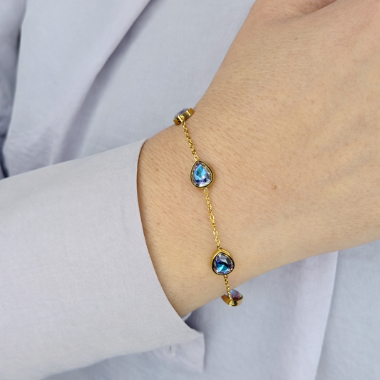 Gouden damesarmband met druppelvormige iriserende zirkonia stenen, gedragen om de pols, close-up productfoto op lichte achtergrond.