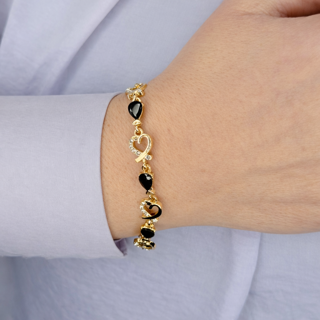 Gouden damesarmband met zwarte hartvormige stenen en hartdetails met fonkelende kristallen, gedragen om de pols op een lichte achtergrond.