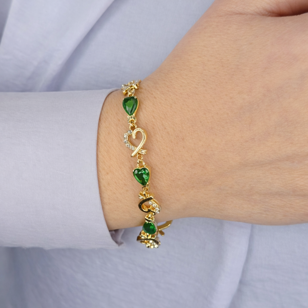 Gouden damesarmband met groene hartvormige stenen en hartdetails met fonkelende kristallen, gedragen om de pols op een lichte achtergrond.
