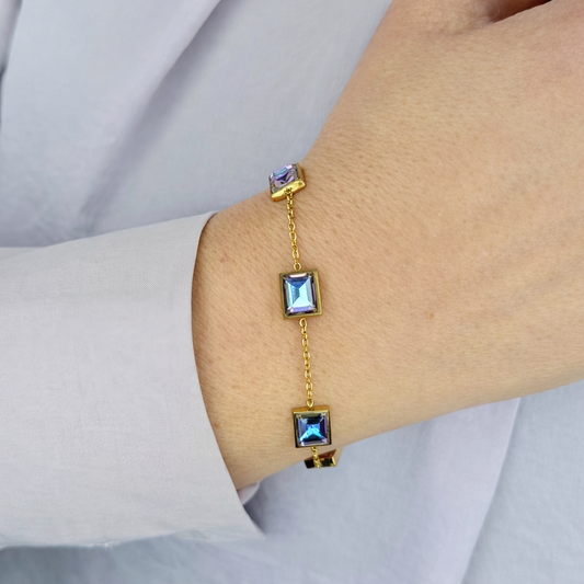 Gouden armband met vier vierkante blauwe zirkonia stenen, gedragen om de pols, close-up productfoto op lichte achtergrond.